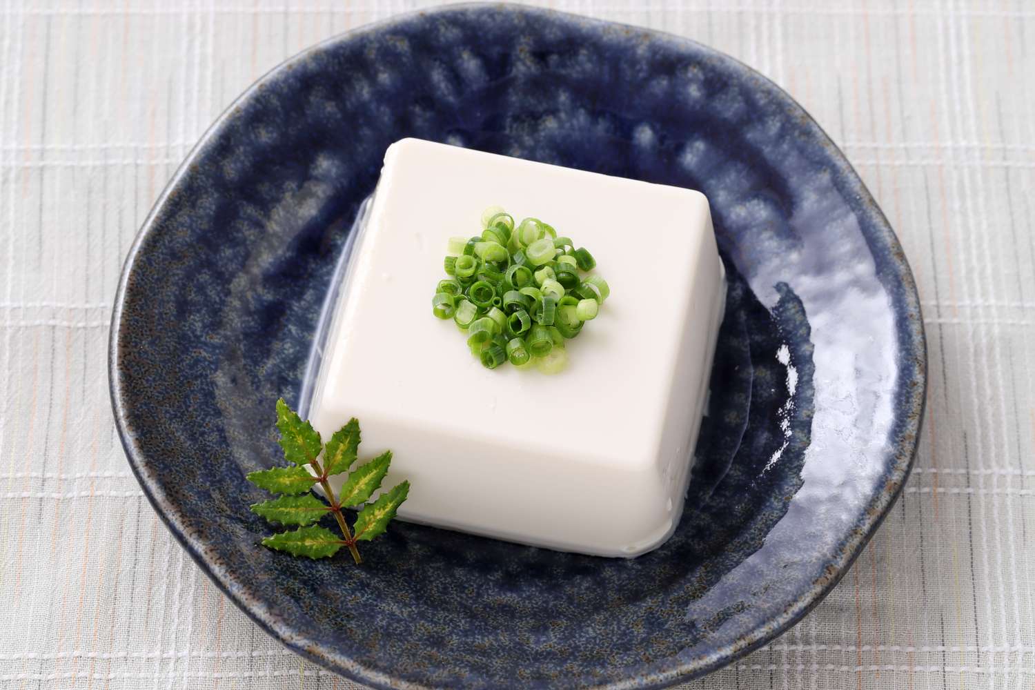 block of silken tofu garnished with chopped scallions in dark blue dish, t
