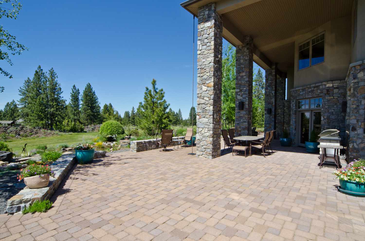A patio area with potted plants and outdoor furniture next to a house, surrounded by trees and a grassy landscape