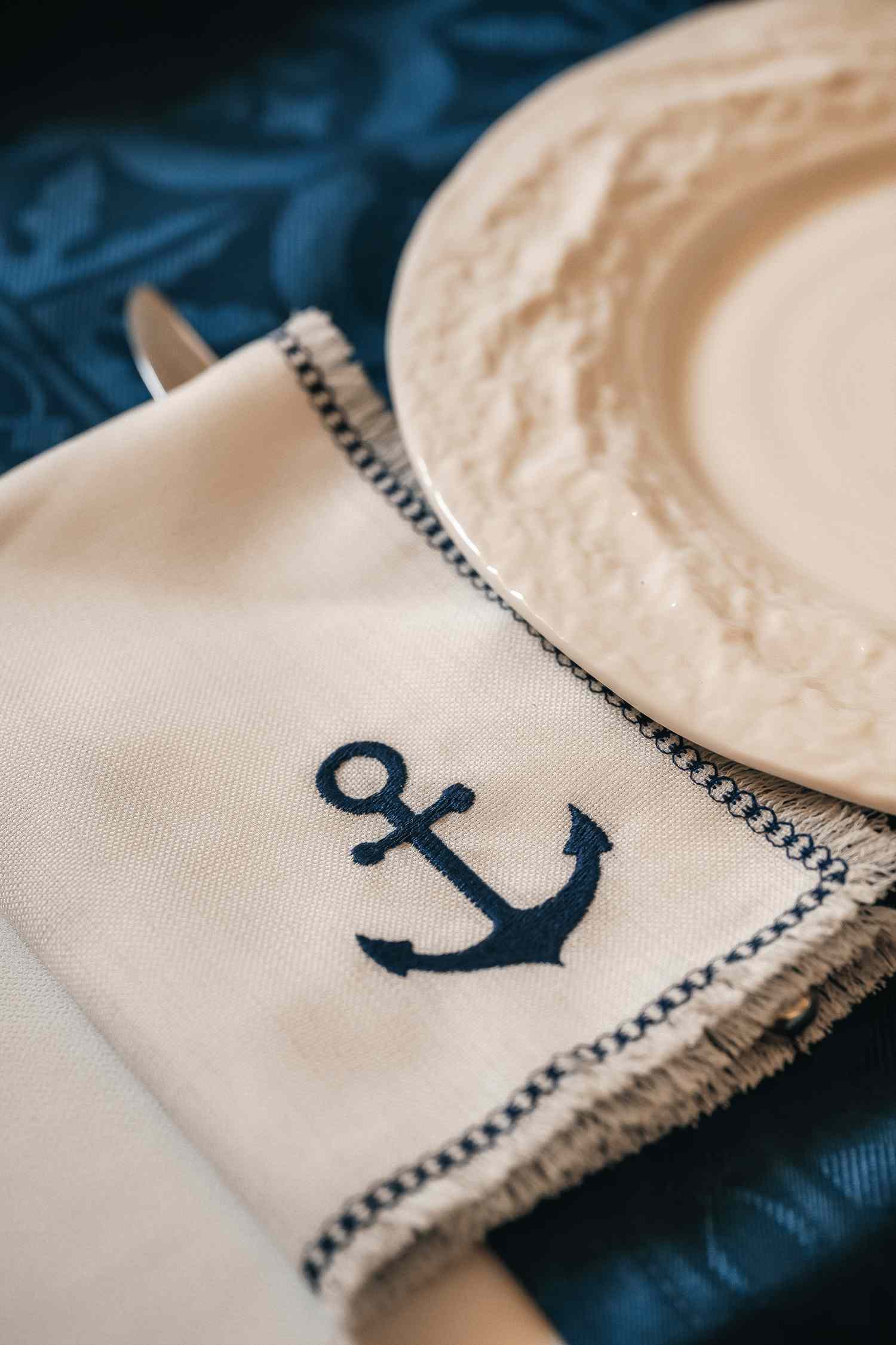 A decorative table setting featuring a napkin embroidered with an anchor design beside a plate