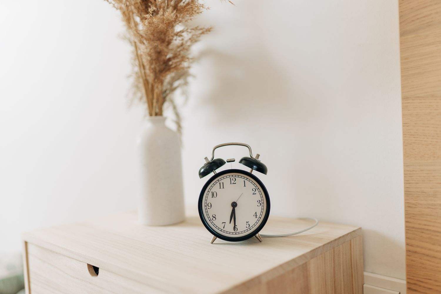An alarm clock on a wooden surface next to a vase with dried plants