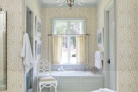 A bathroom with a patterned wallpaper a bathtub centered below a window a chair and neatly arranged towels and artwork