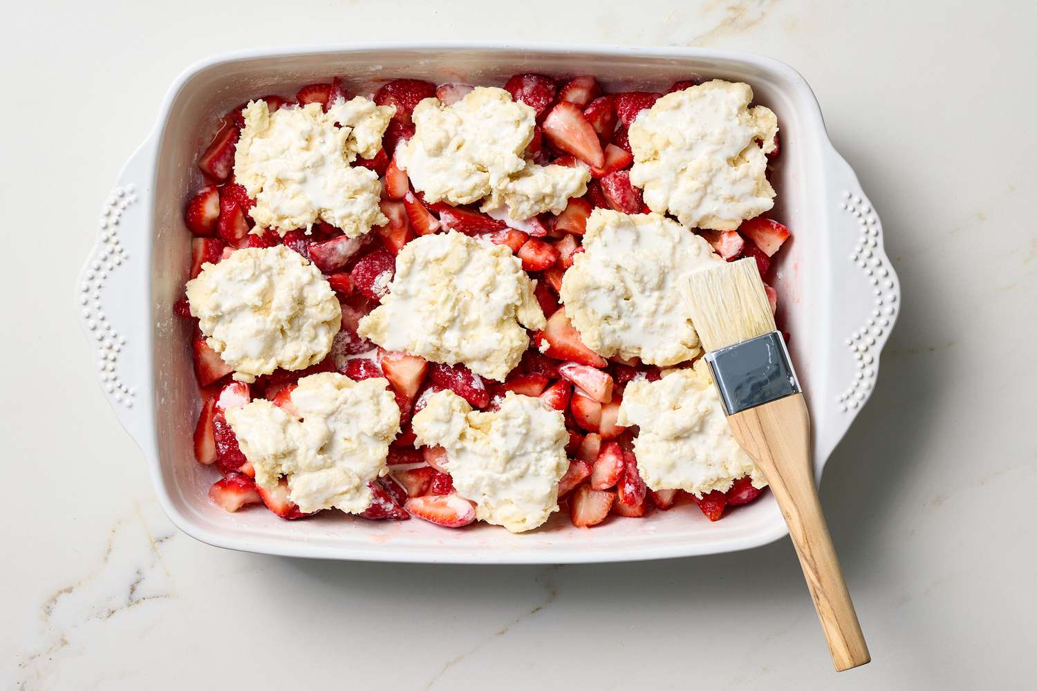 A baking dish with strawberry cobbler ready to be baked, uncooked dough topping over cut strawberries