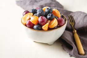 A bowl of assorted fruits, including grapes, blueberries, and mandarin slices, on a table with a fork placed nearby