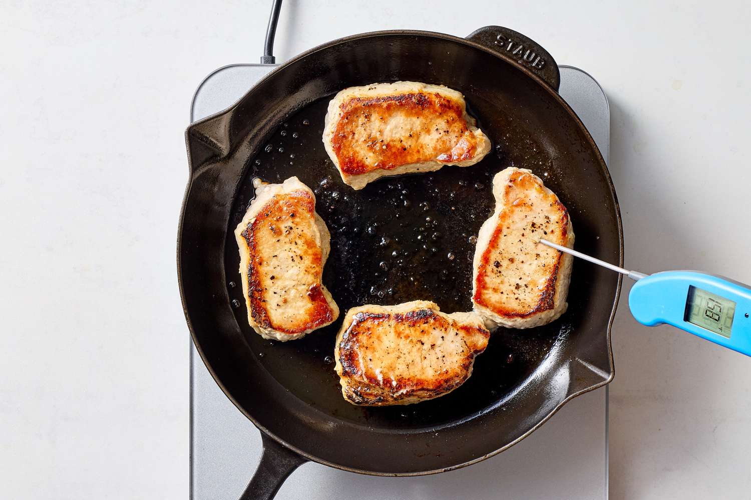 Four pork chops cooking in a skillet with a thermometer checking one chop