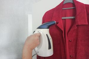A handheld garment steamer in use on a red shirt hanging on a hanger