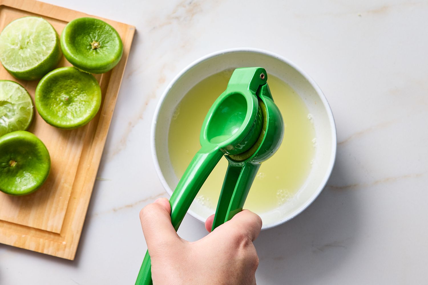 Hand using a citrus squeezer with halved limes and a bowl of juice