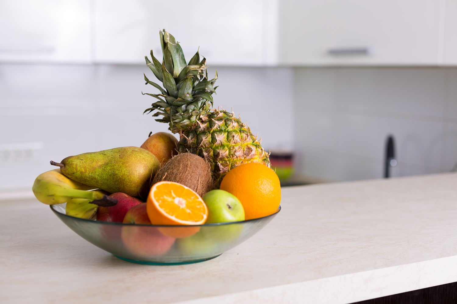A bowl of various fruits including a pineapple, oranges, and pears placed on a kitchen counter