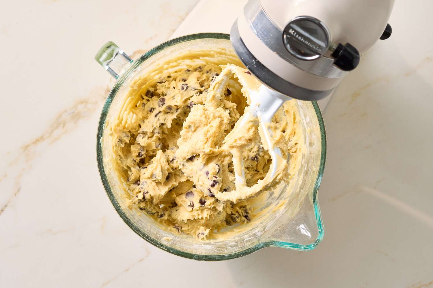 Bowl of cookie dough with chocolate chips and a stand mixer paddle attachment resting on the side