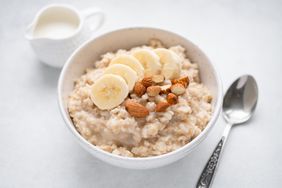 A bowl of oatmeal topped with banana slices and nuts spoon nearby and small pitcher in background