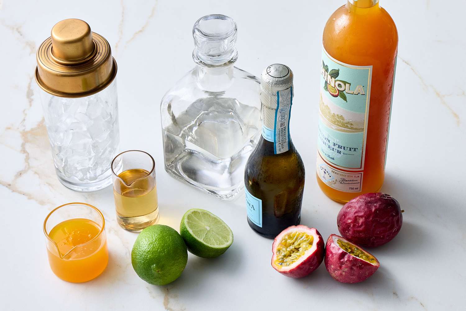 Ingredients for making a passion star martini, including a cocktail shaker, spirits, lime, passionfruit, and mixers on a marble surface