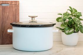 A slow cooker on a kitchen counter with a plant and a wooden cutting board in the background