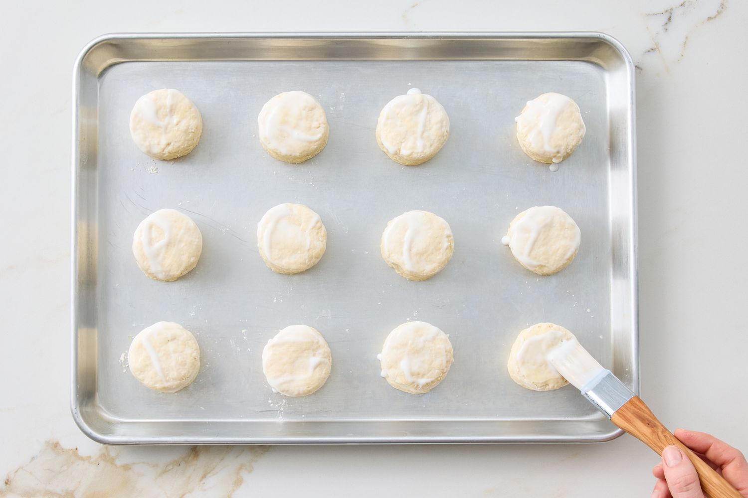 A baking tray with evenly spaced unbaked biscuits a hand brushing one biscuit with liquid using a pastry brush