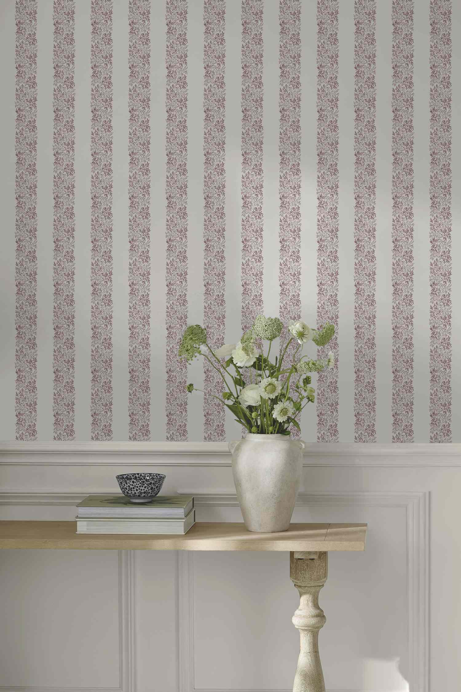 A hallway scene with a small table books a vase with flowers and a striped wallpaper in the background