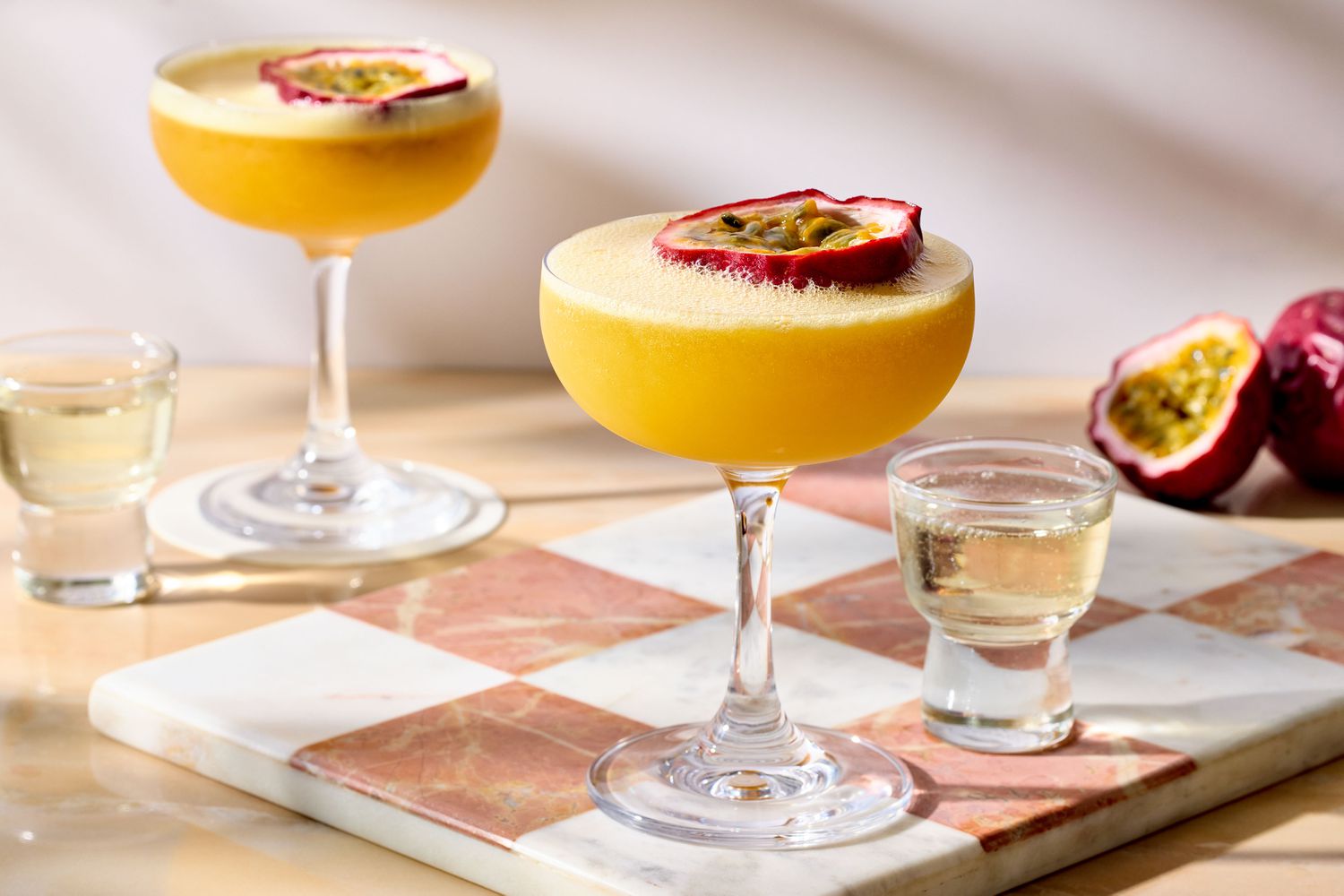 Two passion fruit martinis garnished with passion fruit halves, alongside two small glasses of liquid on a checkered surface