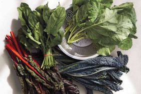 Various fresh leafy greens arranged on a surface including chard spinach kale and collard greens around a colander