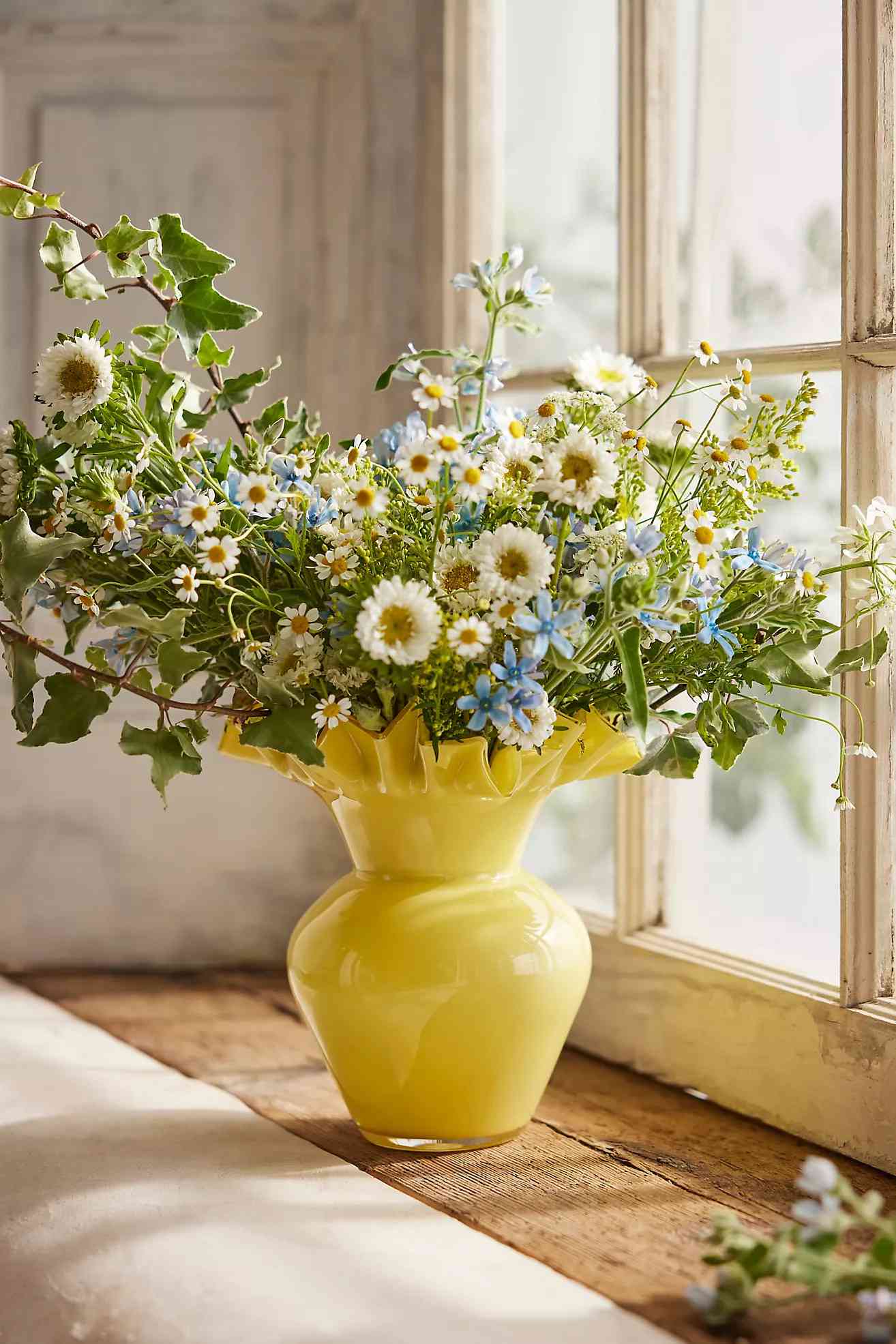 A yellow glass vase holding a bouquet of daisies, blue flowers, and greenery on a wooden surface by a window