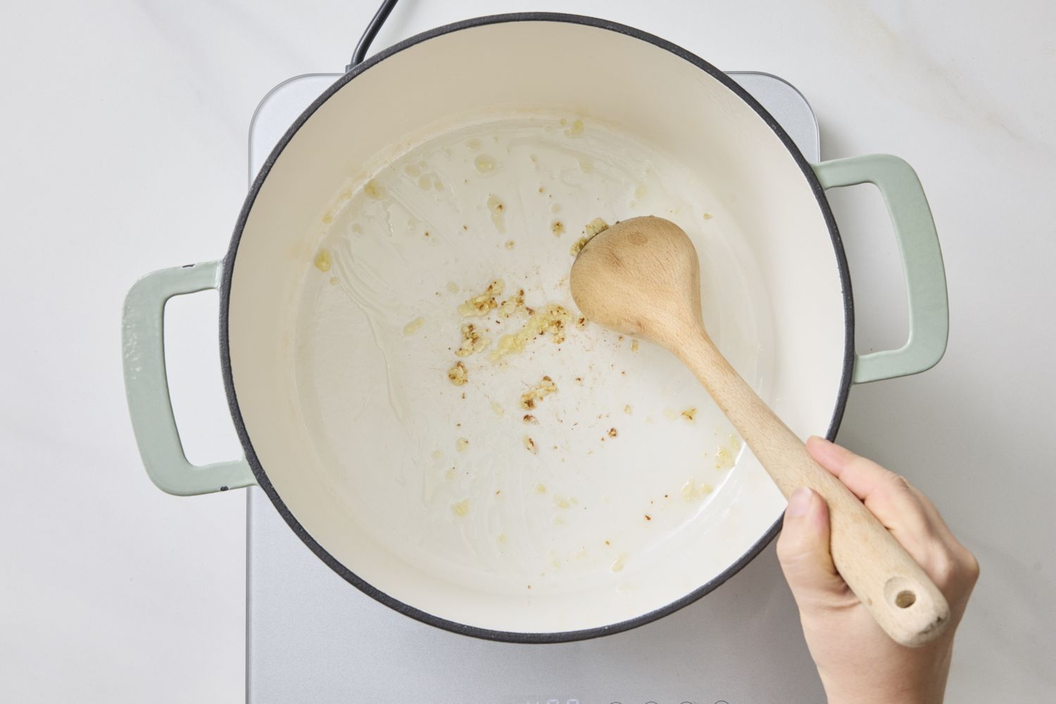 A hand stirring a nearly empty pot with a wooden spoon visible stove beneath
