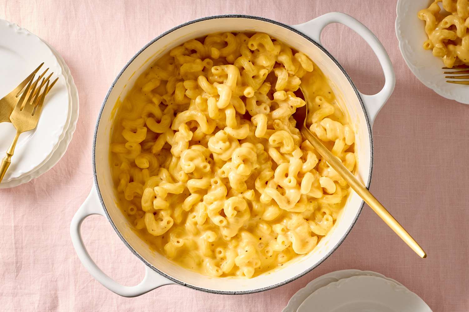 A pot of macaroni and cheese with a serving spoon placed on a table with plates and forks nearby