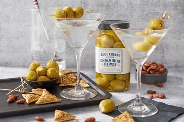 Two martini glasses garnished with olives a jar of blue cheese stuffed olives crackers almonds and a drink in the background