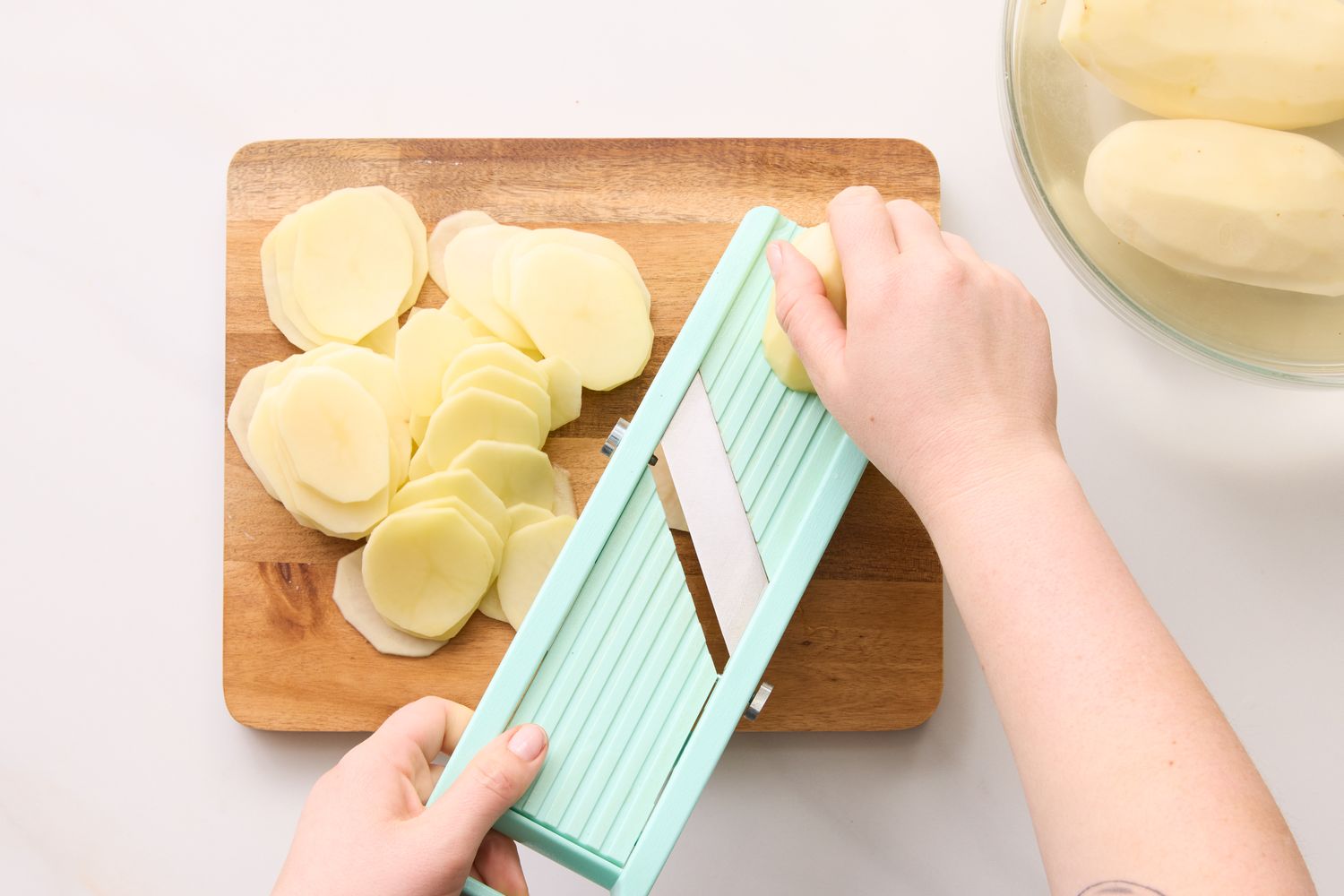 Hands slicing potatoes using a mandoline slicer on a wooden cutting board