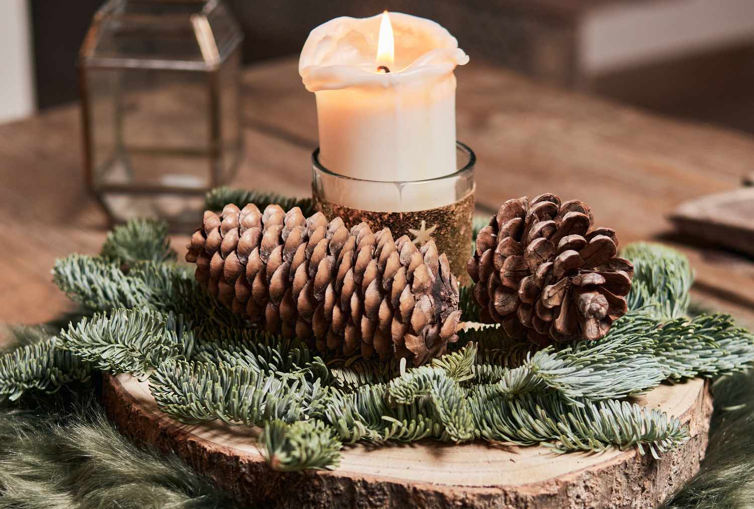 A decorative pinecone arrangement with evergreen branches on a wood slice featuring a lit white candle in a holder