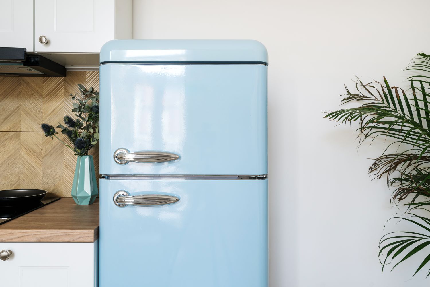 Detail in kitchen interior, blue refrigerator with stainless steel handles in retro style near cabinet on white wall background. Fridge for storage food products at home. Household appliances concept