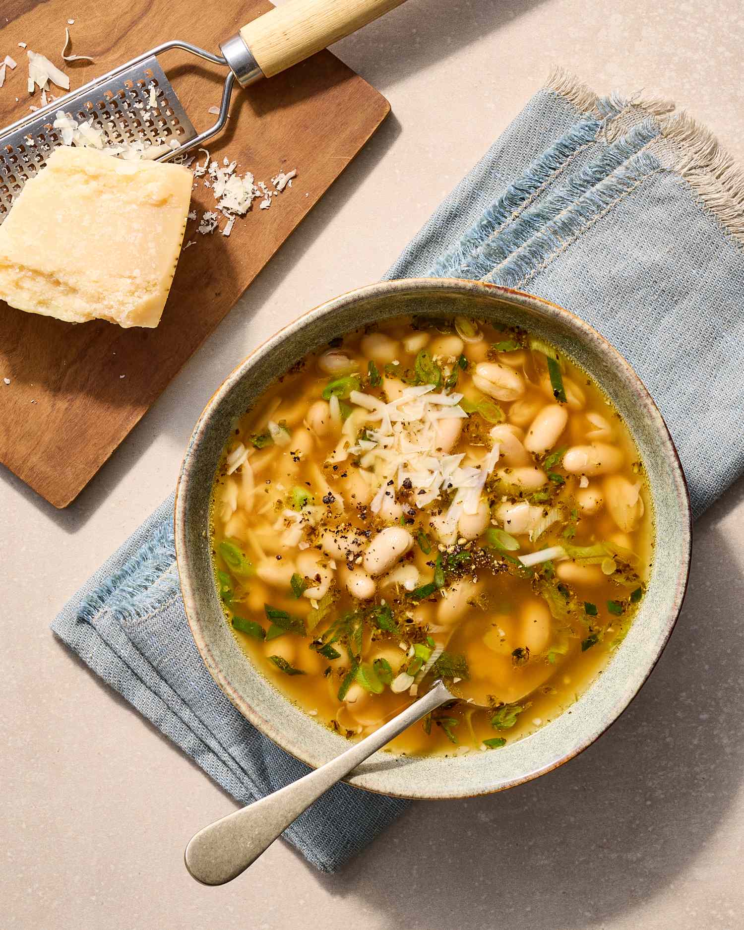 A bowl of white bean soup garnished with herbs next to a wooden board with grated cheese and a grater