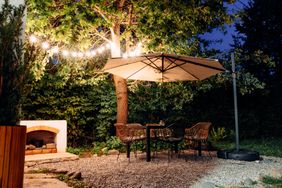 Outdoor patio with chairs around a table under an umbrella, near a tree and illuminated string lights