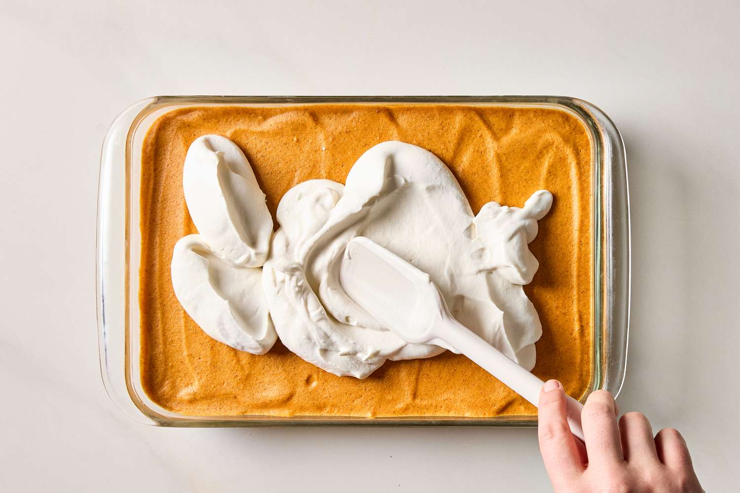 Someone spreading whipped topping onto a pumpkin mousse cheesecake with a spatula in a glass dish