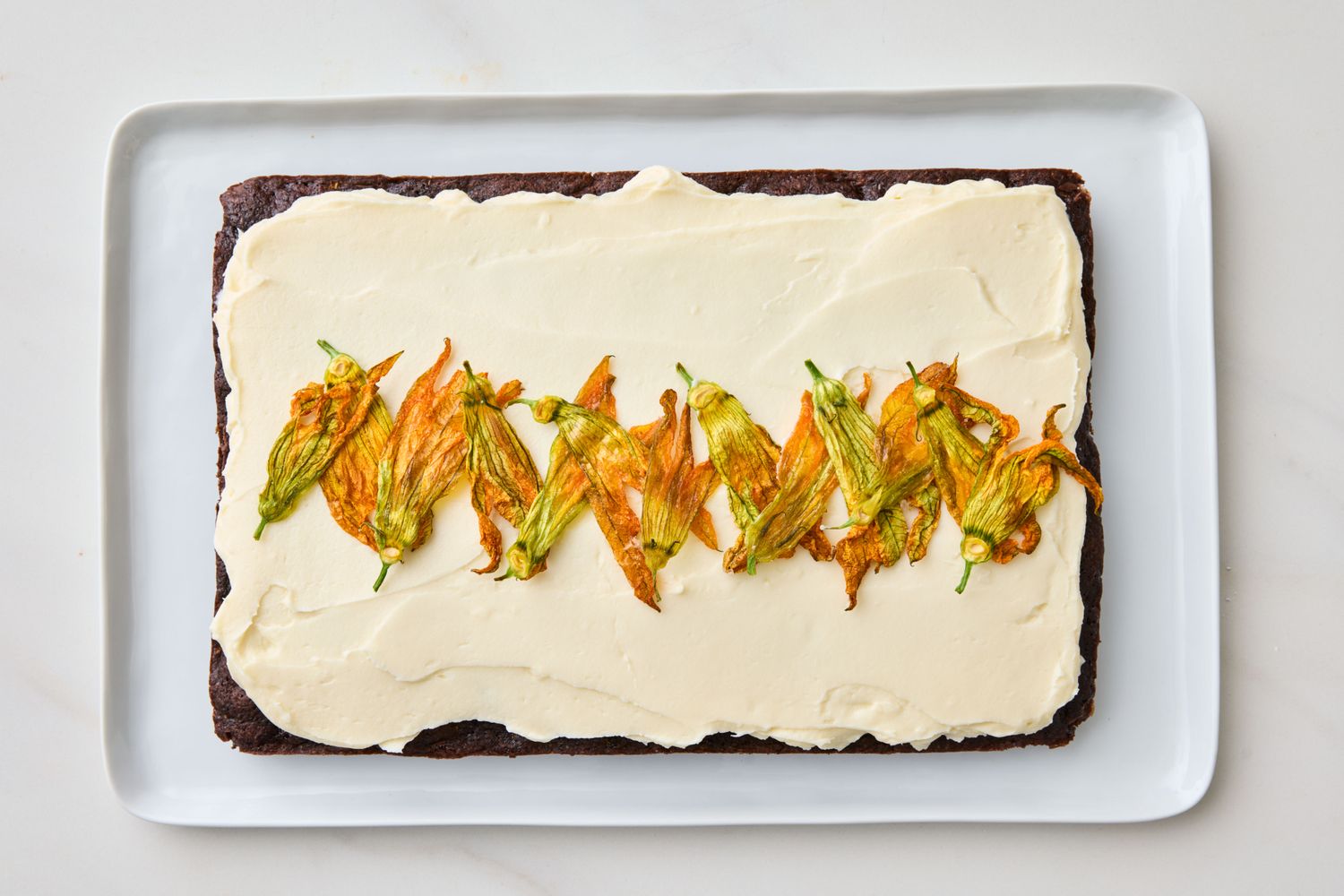 Sheet cake with cream cheese frosting and zucchini flowers on top served on a rectangular platter