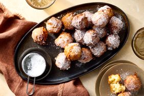 A platter of apple fritters dusted with powdered sugar with a small container of sugar and additional fritters on a side plate
