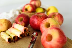 Apples with cores removed placed on a surface a coring tool visible with additional apples and a basket in the background