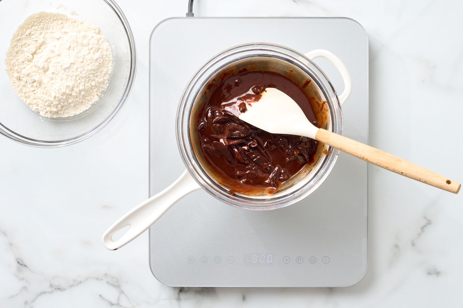 Melted chocolate in a glass bowl with a spatula on an electric cooktop a bowl of flour nearby