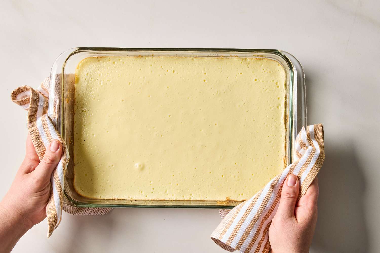 A person holding a rectangular glass pan containing a baked cheesecake using a striped kitchen towel