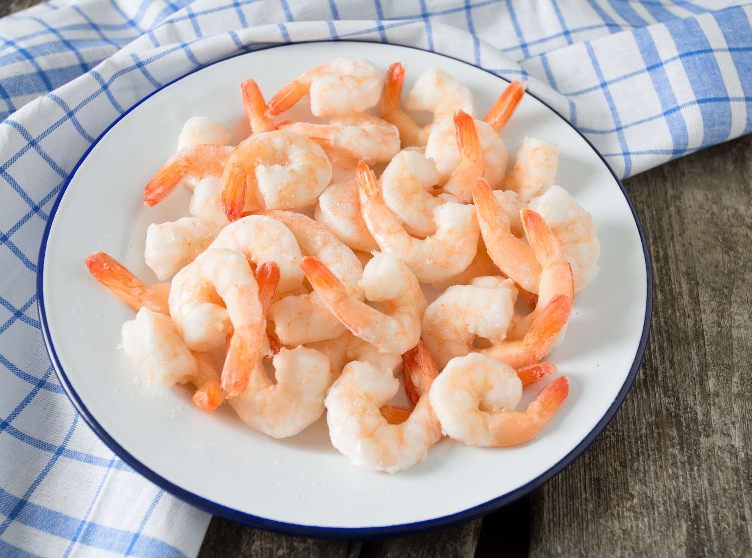 A plate of cooked shrimp with tails arranged on a white dish on a surface with a checkered cloth