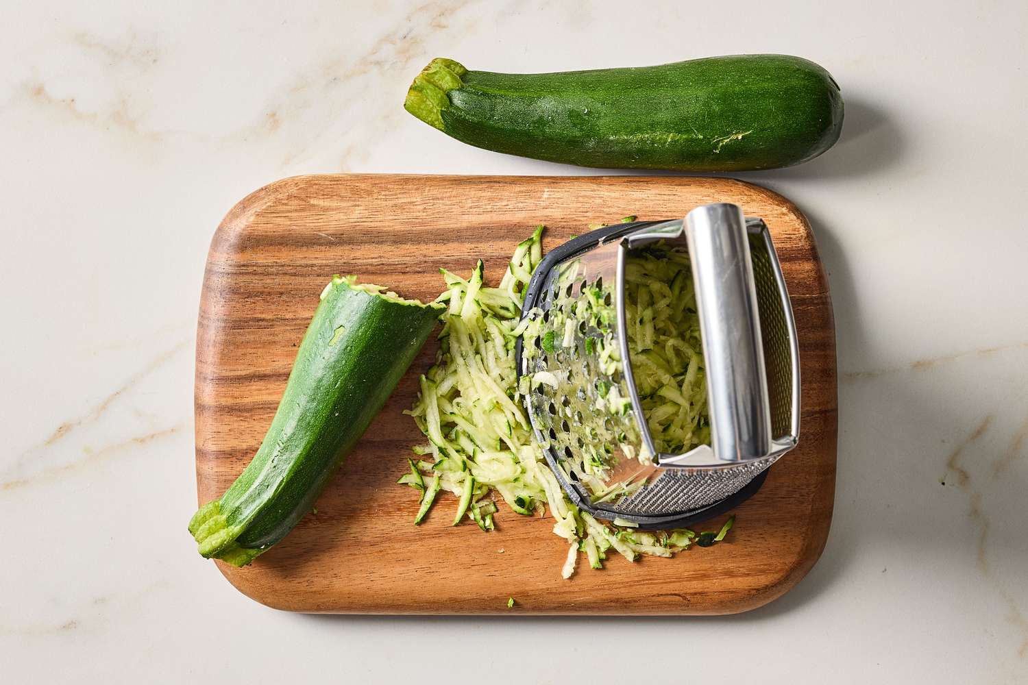 Grated zucchini on a cutting board with a box grater and whole zucchini