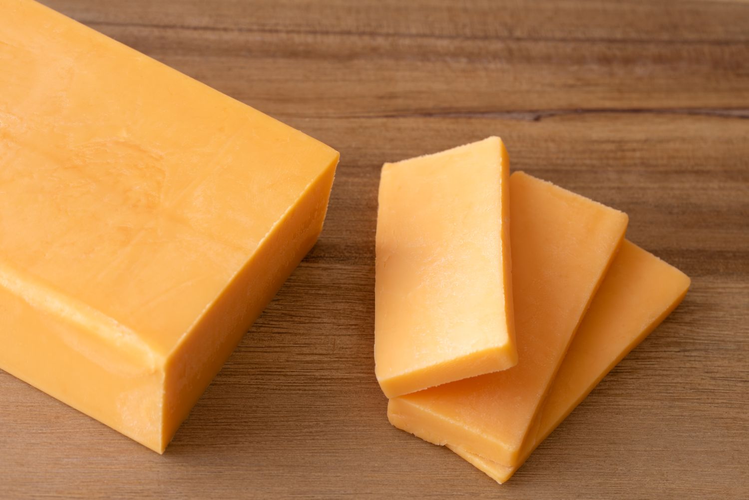 A block of cheddar cheese with three slices placed beside it on a wooden surface