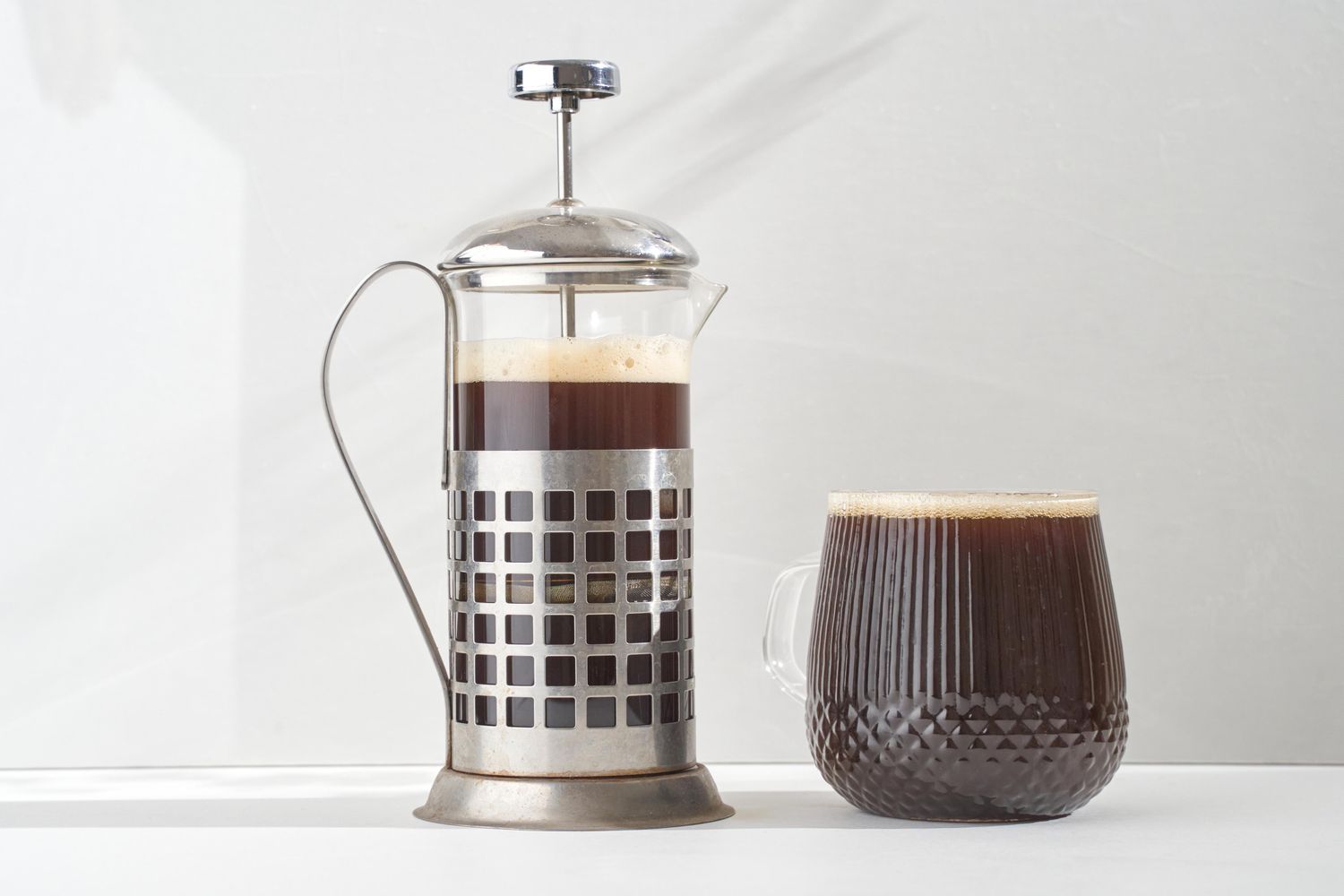 A French press next to a full glass of coffee placed on a white surface