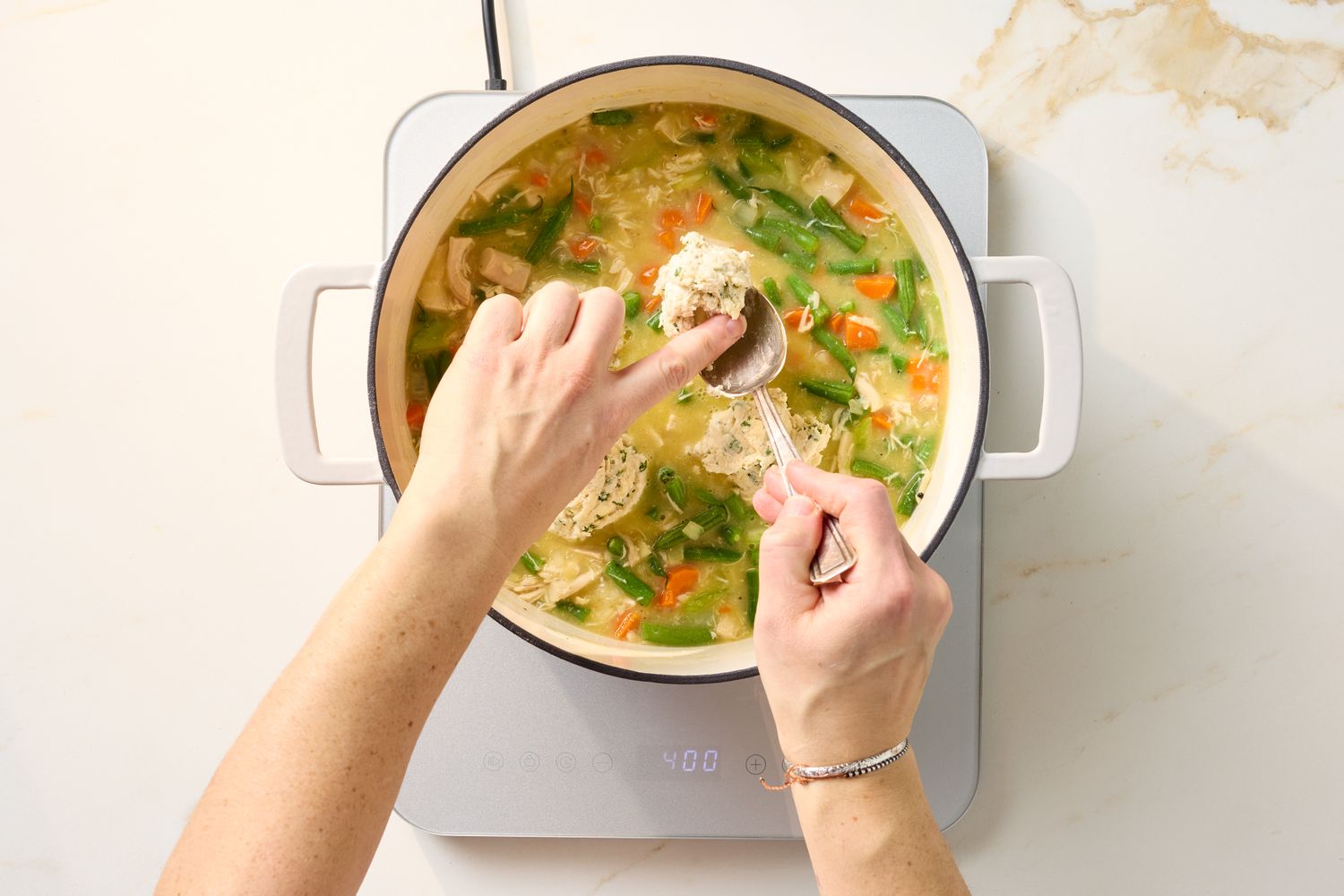 Hands serving chicken and dumplings from a pot on a stovetop