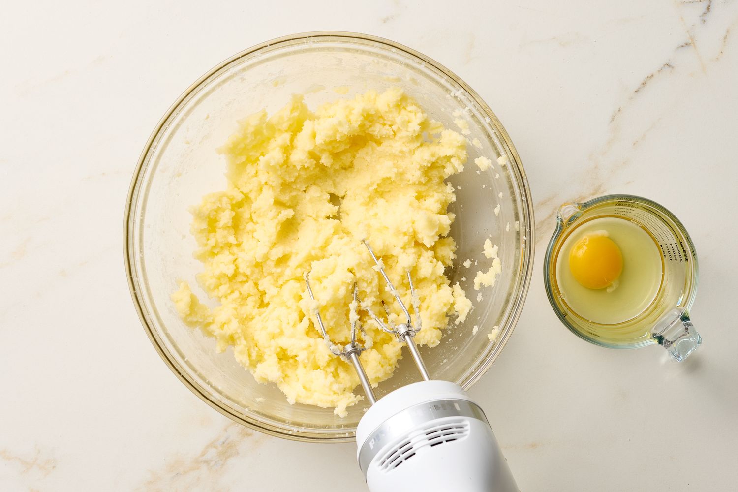 A bowl of sugar cookie dough being mixed with a hand mixer next to a glass container holding an egg