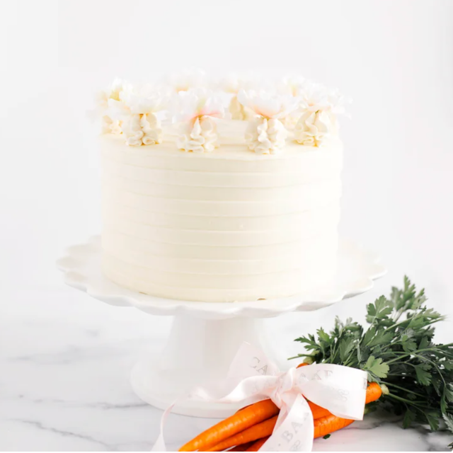 A carrot cake on a white platter decorated with frosting and flowers fresh carrots with leafy tops placed beside it