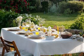 A dining table set outdoors in a garden with plates glasses and floral arrangements