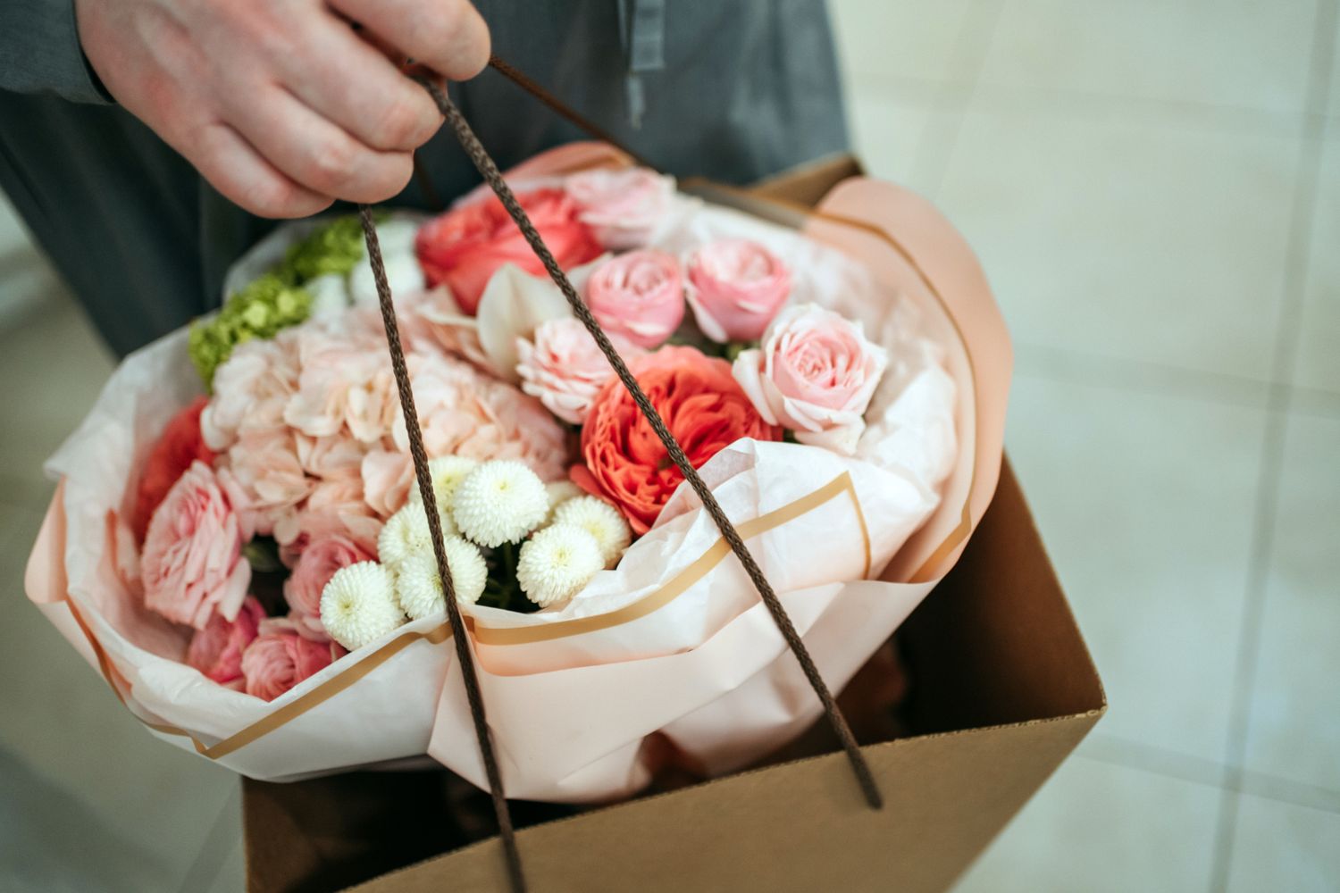 Hand holding a gift bag containing a bouquet of flowers including roses and other blossoms wrapped in paper