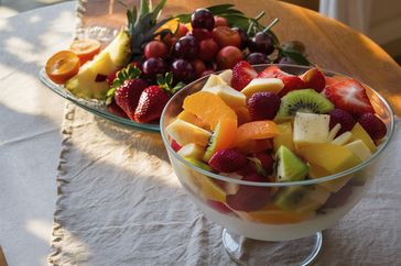 A bowl of fruit salad with kiwi mango orange berries and pineapple with an additional platter of fresh fruit in the background