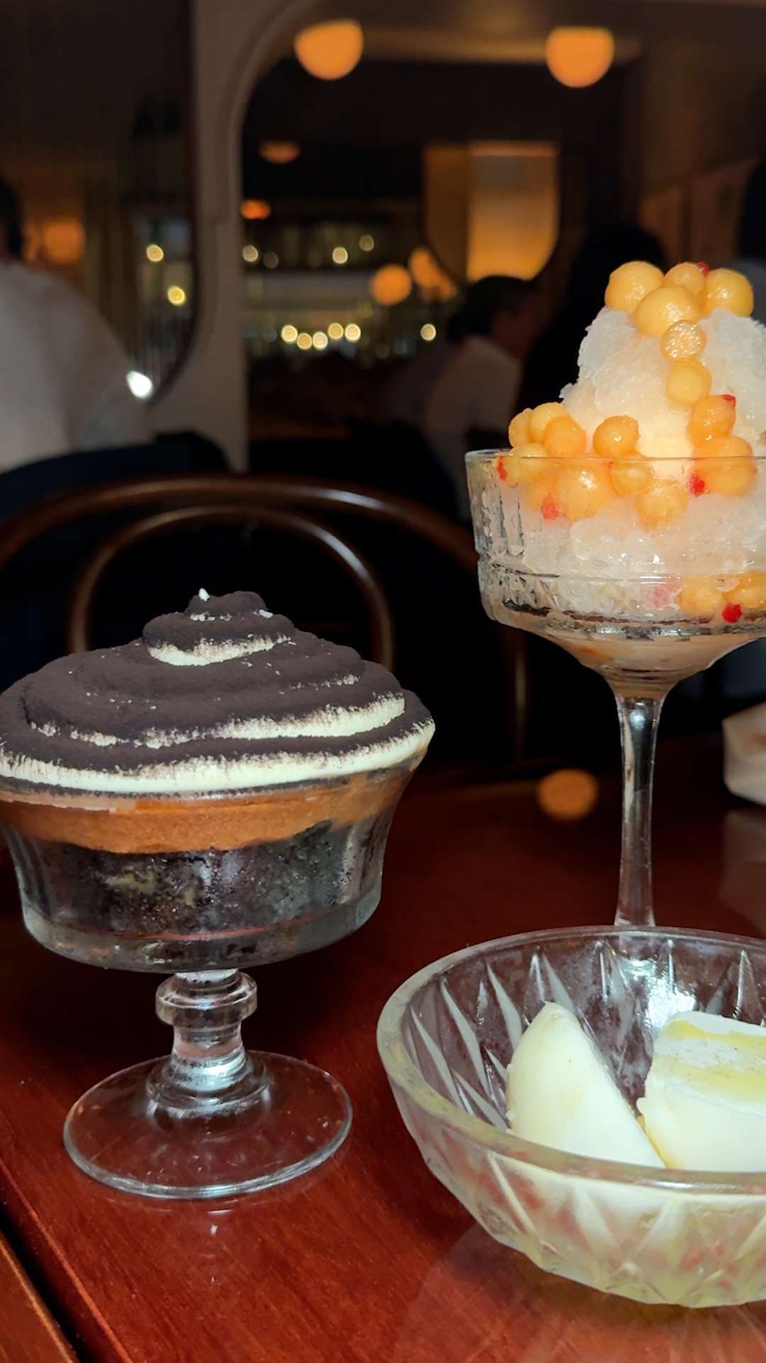 Two desserts in glasses on a table left with chocolate and cream layers right with fruit toppings and ice with a small dish in front