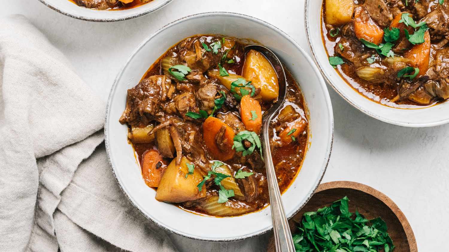 Bowls of stew with vegetables and pieces of meat, garnished with herbs, a napkin and a bowl of chopped greens beside