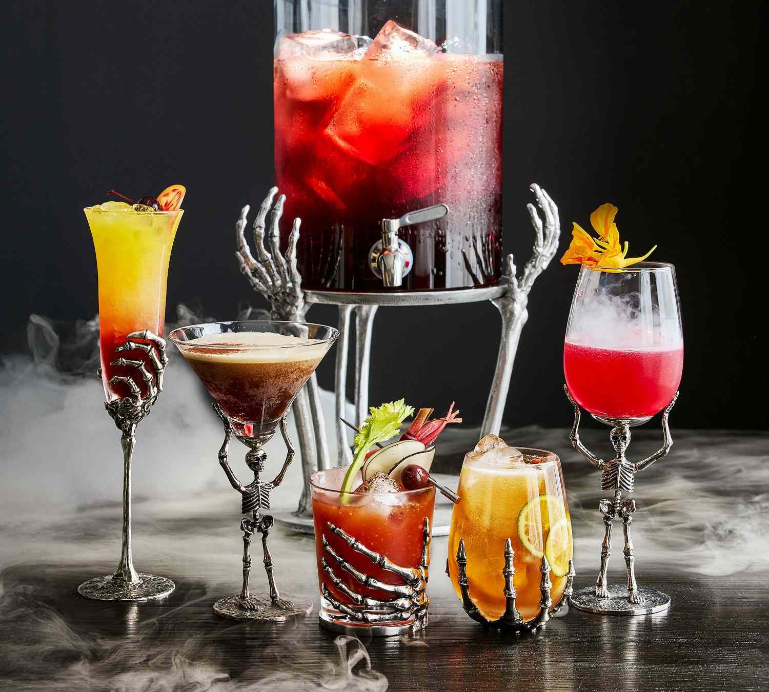 A collection of drinkware designed with skeleton hand motifs holding various cocktails and a red liquid dispenser elevated on skeletal hands
