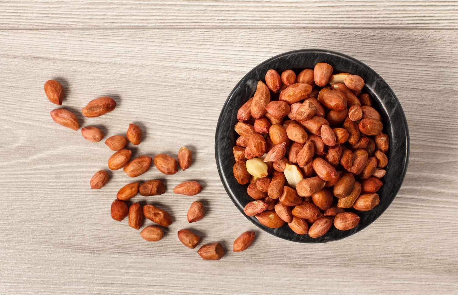 Top view of peanuts in black porcelain bowl and beside it on grey wooden background
