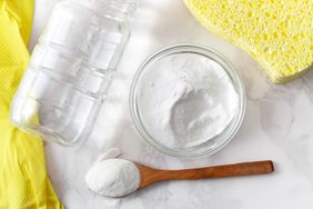 A glass jar a bowl and a wooden spoon with white powder next to a yellow sponge and gloves on a surface
