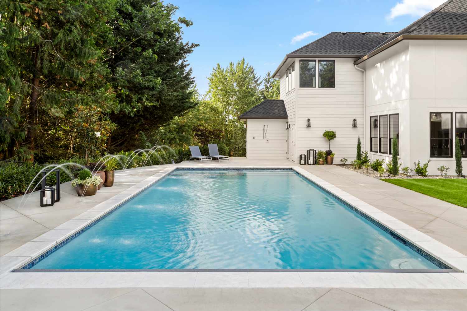 A backyard view featuring a rectangular swimming pool loungers at one end trees and a modern white house on one side